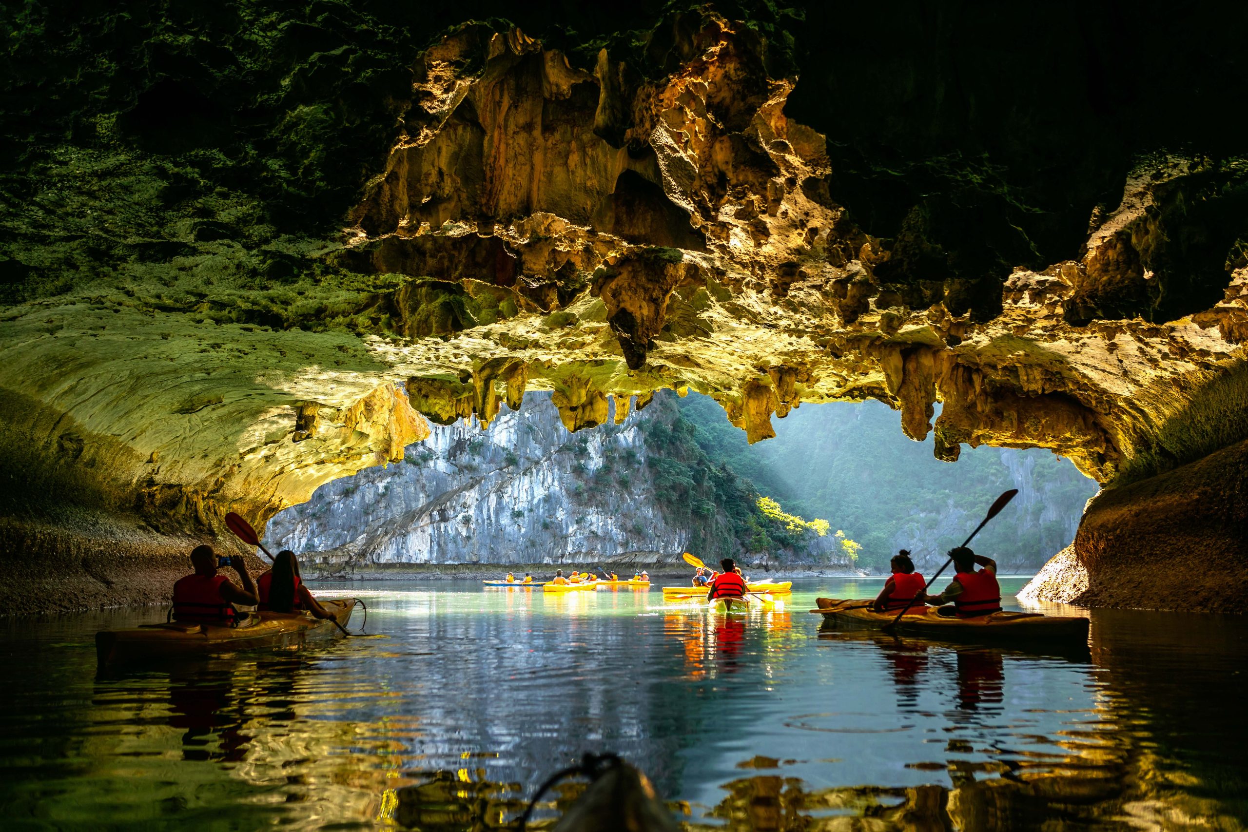Kayakers paddling into a lit limestone cave passage in Lan Ha Bay