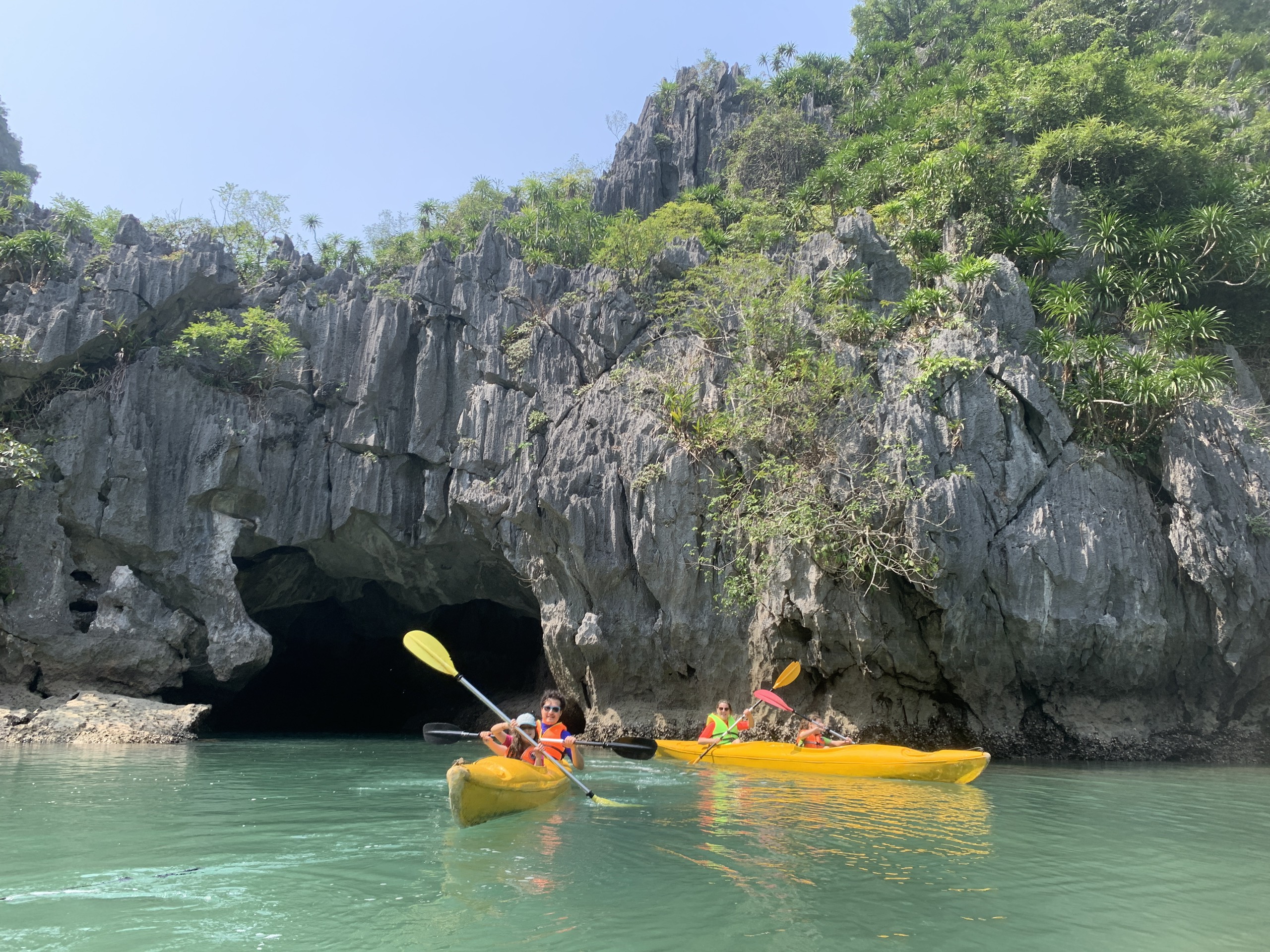 Kayakers paddling past towering limestone cliffs in calm green water, Lan Ha Bay
