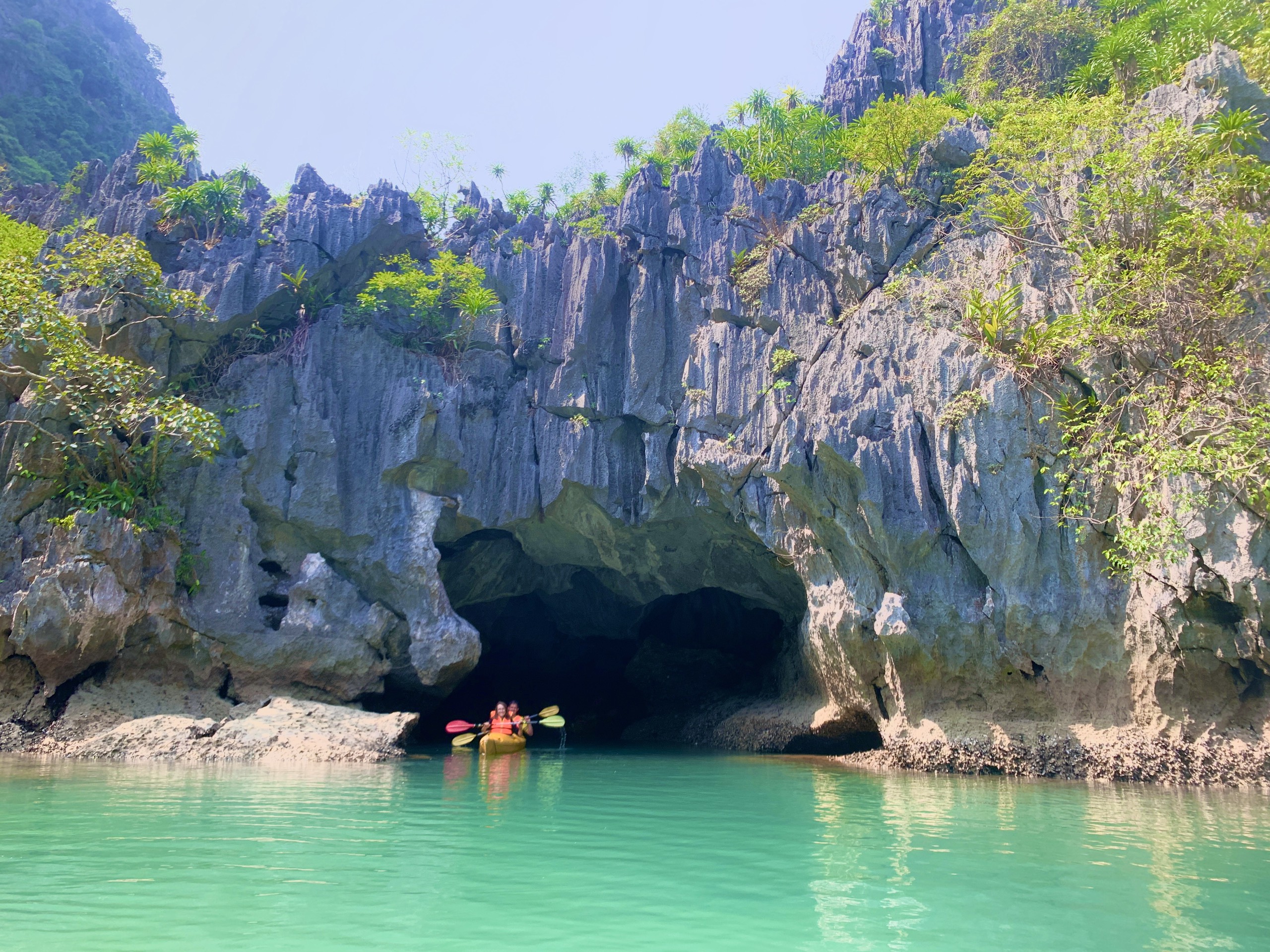 Kayakers paddling into a low natural cave tunnel through a limestone cliff, Lan Ha Bay