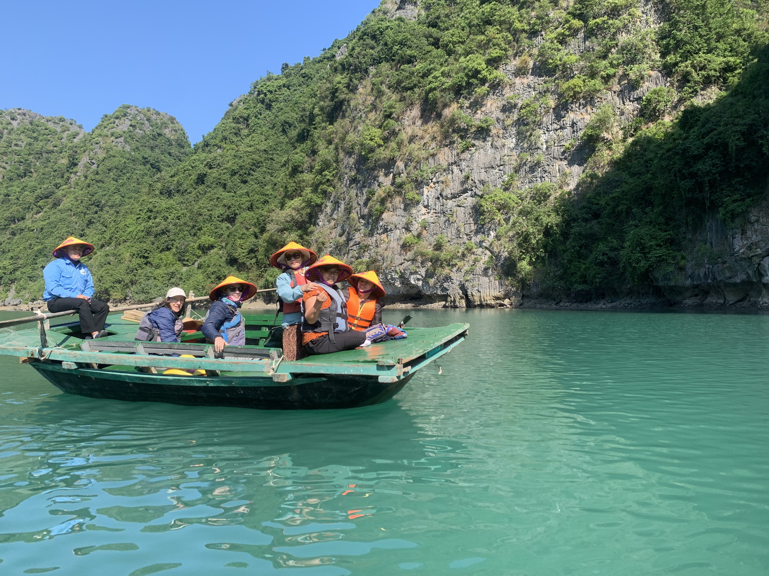 Travellers on a small wooden boat crossing green water between karst islands near Cat Ba
