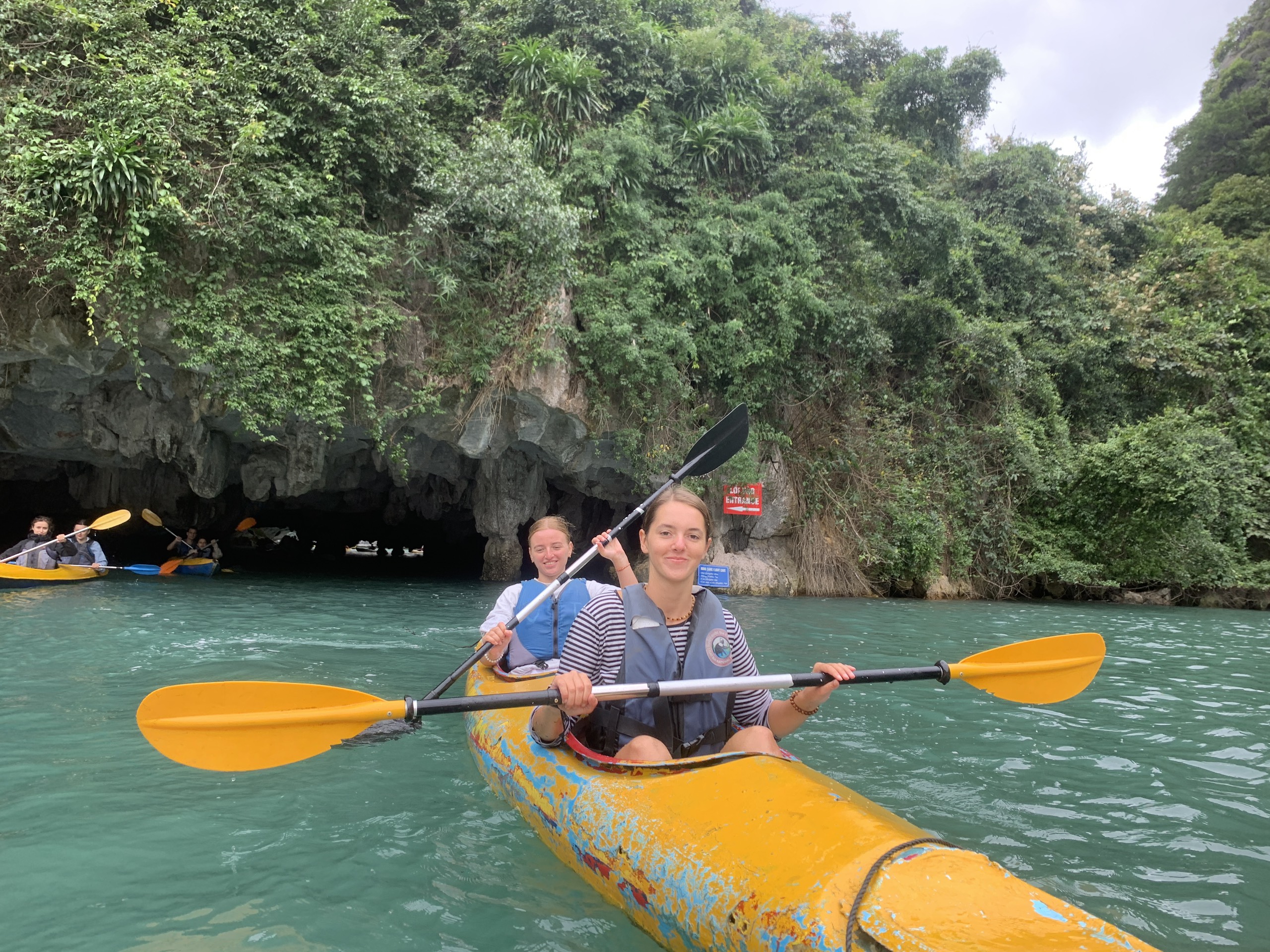 Two people kayaking with orange paddles near a limestone cliff in Lan Ha Bay
